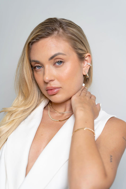 A blonde woman wearing sparkling gold jewelry, including layered necklaces, hoop earrings, and a cuff, gently touches her neck.