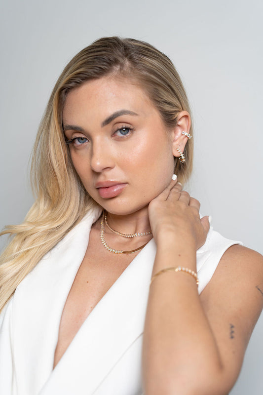 A blonde woman wearing sparkling gold jewelry, including layered necklaces, a bracelet, an earring, and an ear cuff, poses against a neutral background.