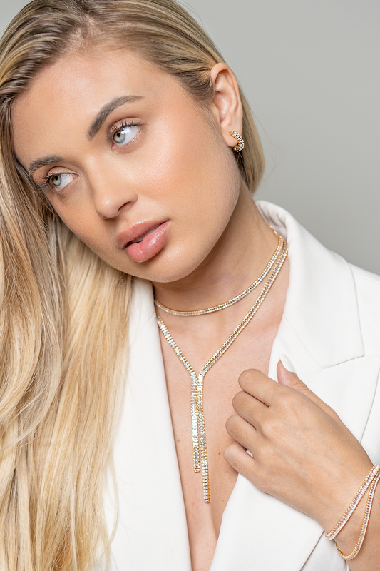 A blonde woman wearing a white blazer showcases sparkling gold jewelry, including layered diamond tennis necklaces, matching hoop earrings, and a tennis bracelet.