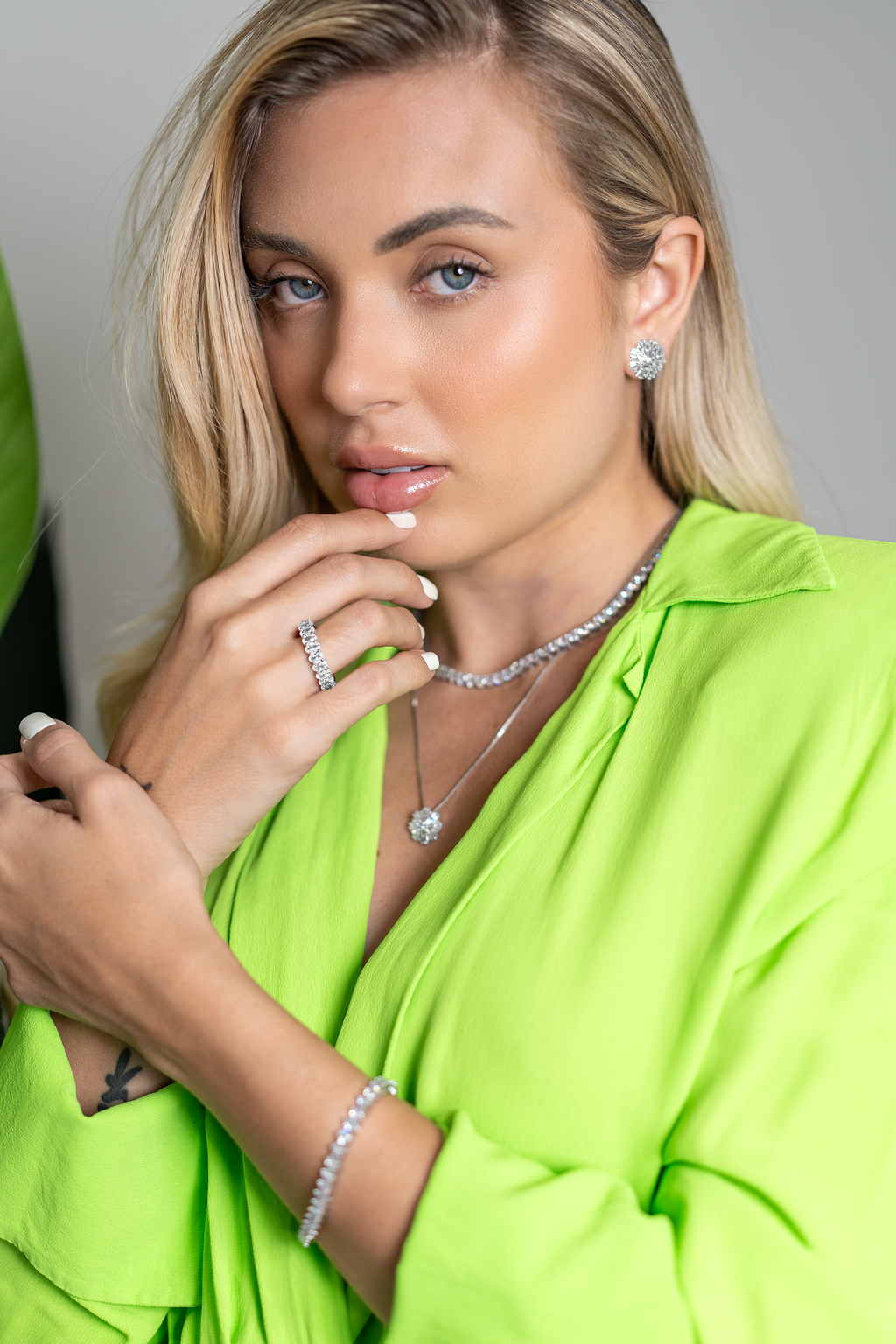 A blonde woman wearing a vibrant green shirt gently touches her lips while showcasing sparkling diamond jewelry, including a tennis necklace, matching earrings, a tennis bracelet, and an eternity ring.