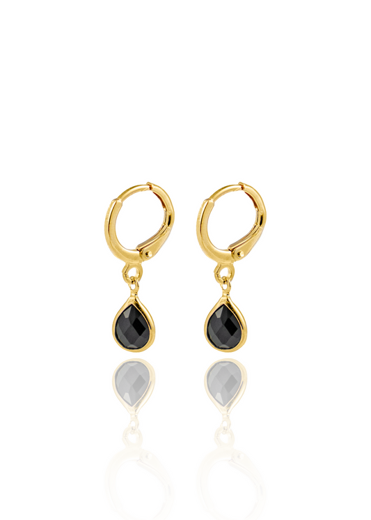 A pair of gold hoop earrings with dangling black teardrop gems gleams against a white background with a soft reflection below.