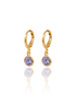 A pair of elegant gold huggie hoop earrings features dangling round purple gemstone charms reflecting on a white surface.