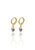 A pair of gold huggie hoop earrings featuring dangling purple heart-shaped gemstones reflects on a white surface.