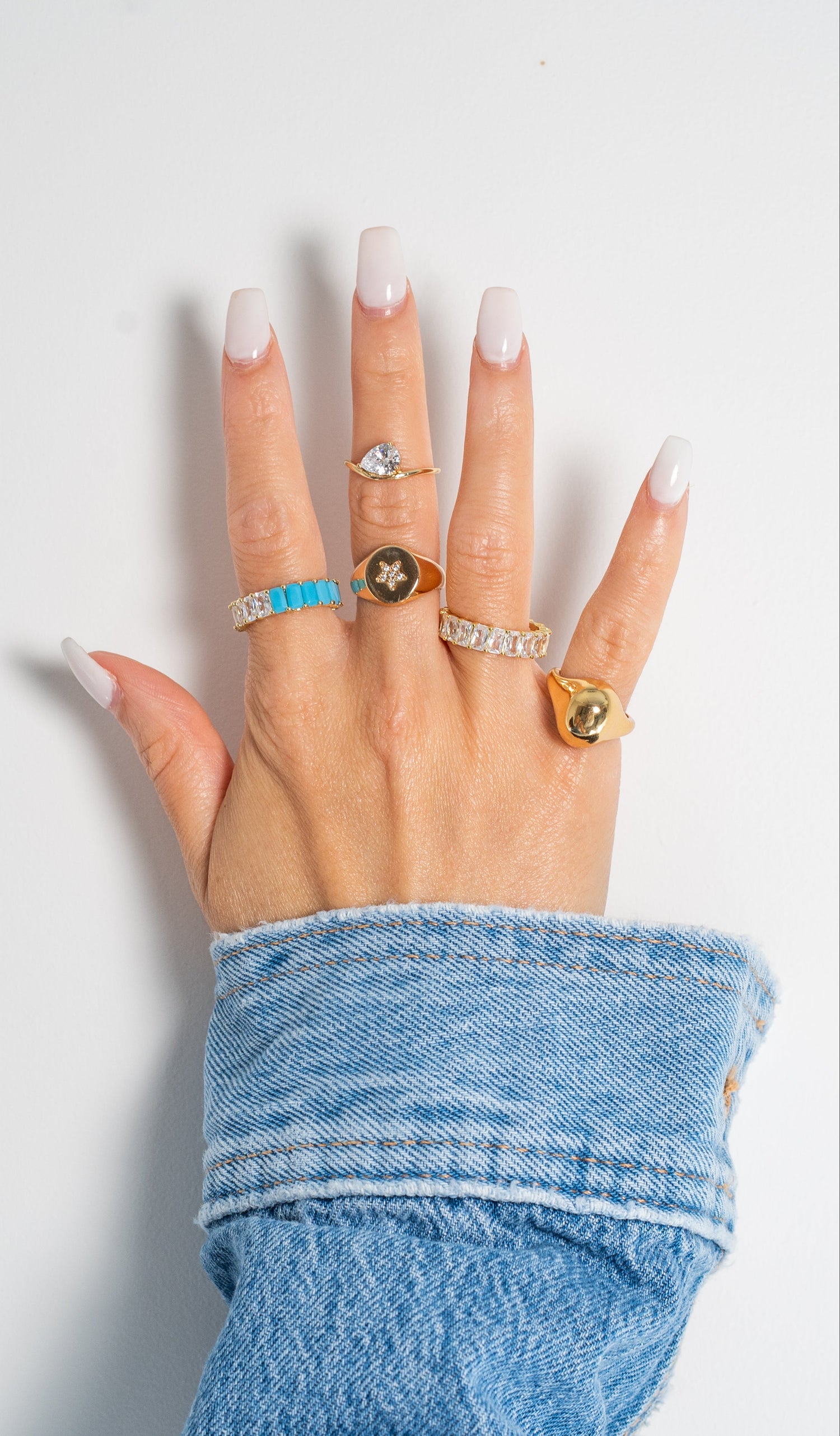 A hand with long white manicured nails wearing five distinct rings, including gold rings with turquoise, star, and oval green stones, and a silver band with clear stones. The hand is resting on a light gray surface with a blue denim sleeve visible.