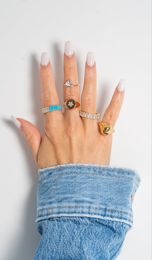 A hand with long white manicured nails wearing five distinct rings, including gold rings with turquoise, star, and oval green stones, and a silver band with clear stones. The hand is resting on a light gray surface with a blue denim sleeve visible.