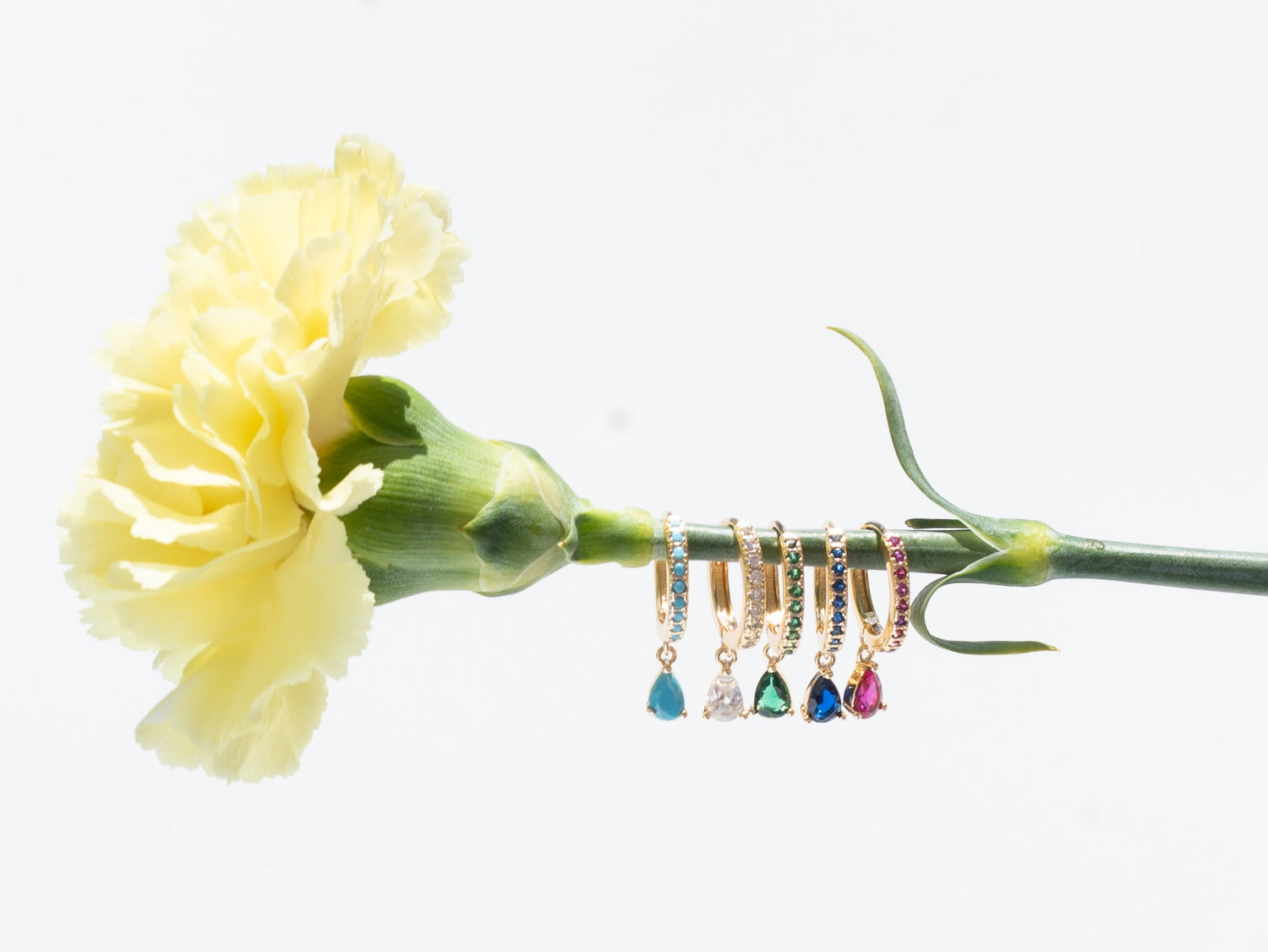 Five small dangling earrings with colorful teardrop-shaped stones in gold-toned settings, hanging from a green flower stem against a plain white background.