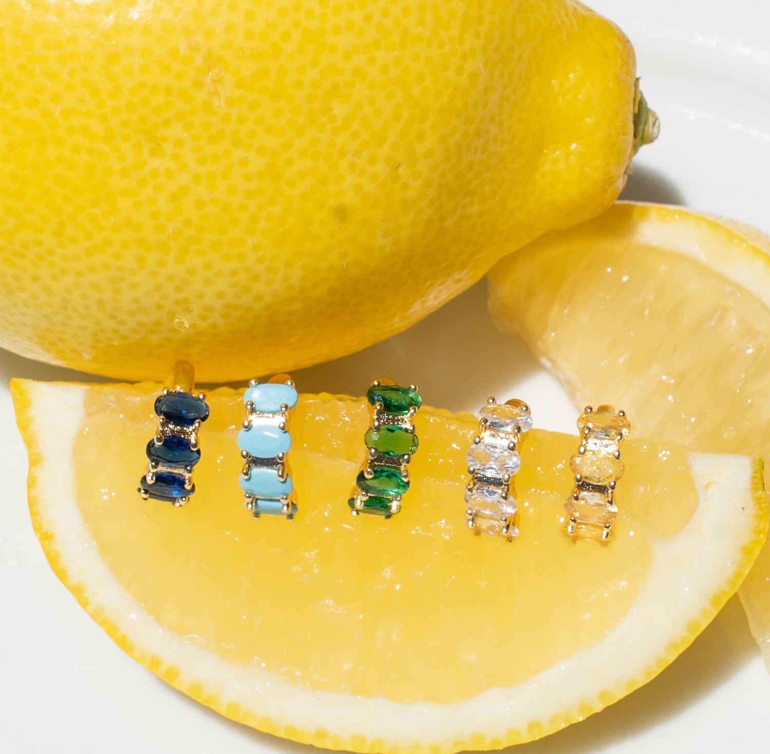 Six pairs of rectangular gemstone EAR CUFFS in various colors (blue, light blue, green, clear, gold) are lined up on a lemon slice, with a whole lemon and another lemon wedge in the background on a white surface.