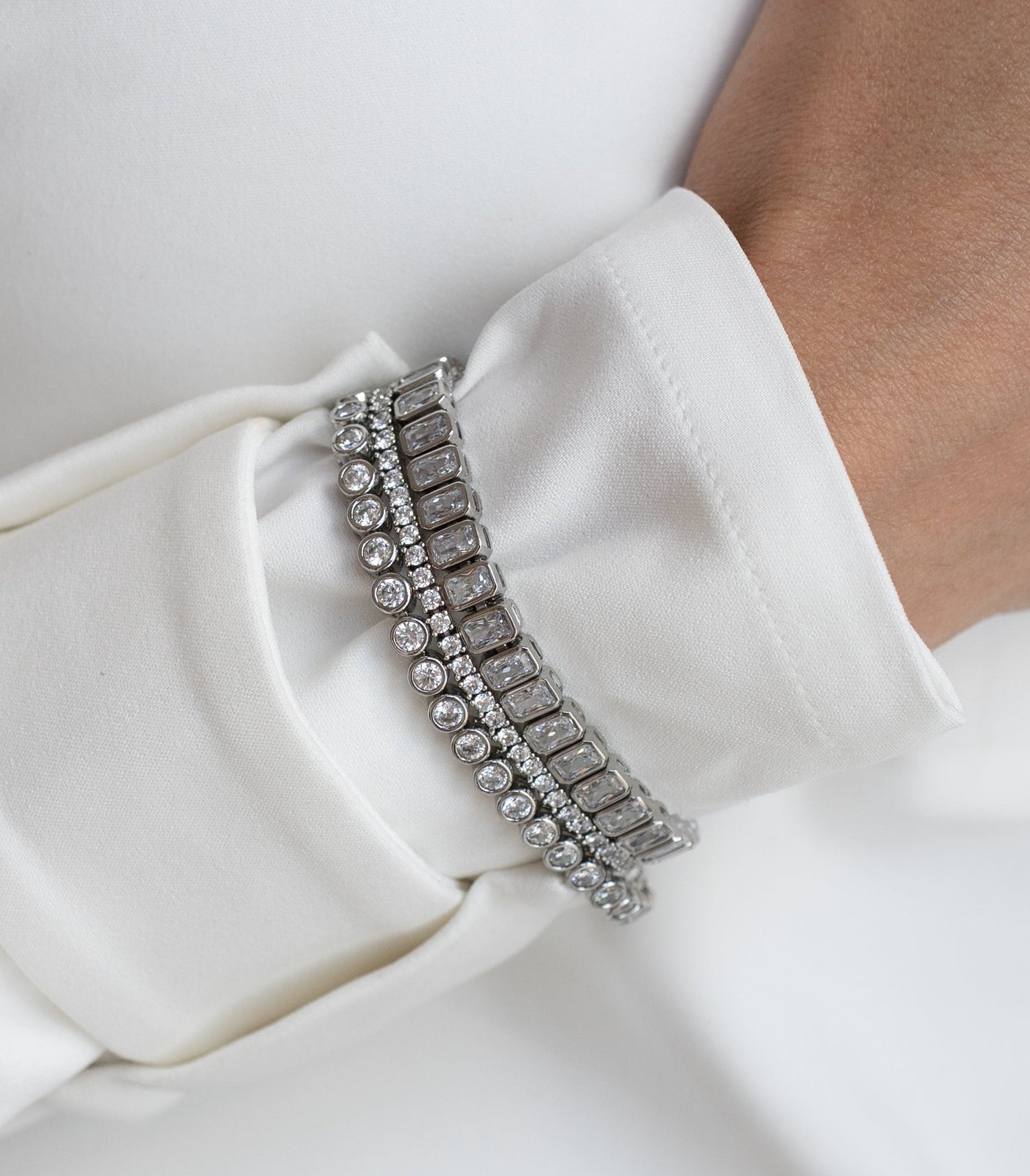 A close-up of a person's wrist wearing a double-row silver bracelet embellished with clear rectangular gemstones, styled with a white satin long-sleeve top.