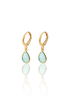 A pair of gold huggie hoop earrings featuring dangling teardrop-shaped light blue gemstone charms rests on a white reflective surface.