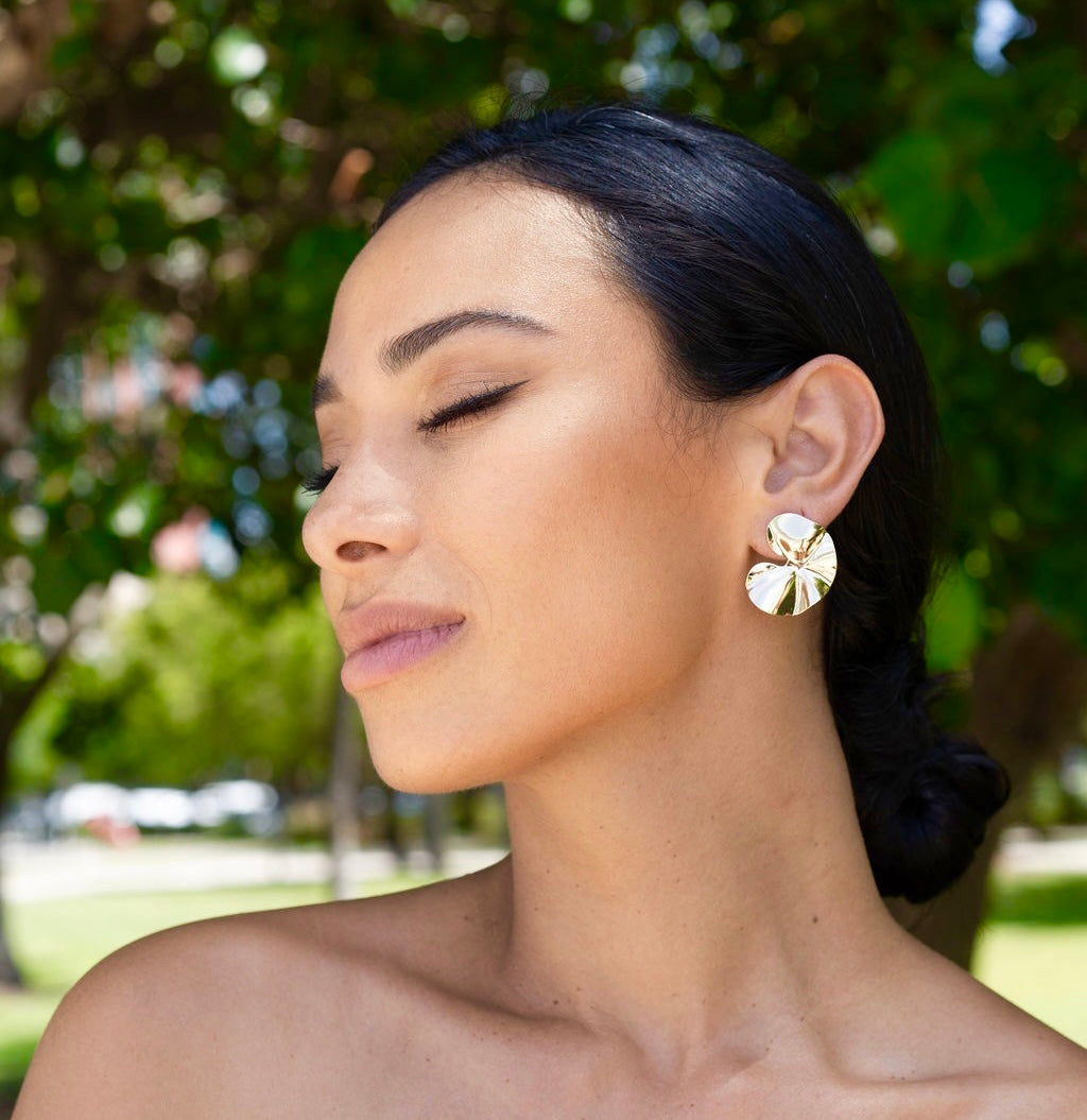 A woman with dark hair tied back showcases sculptural gold statement earrings against a leafy green background.