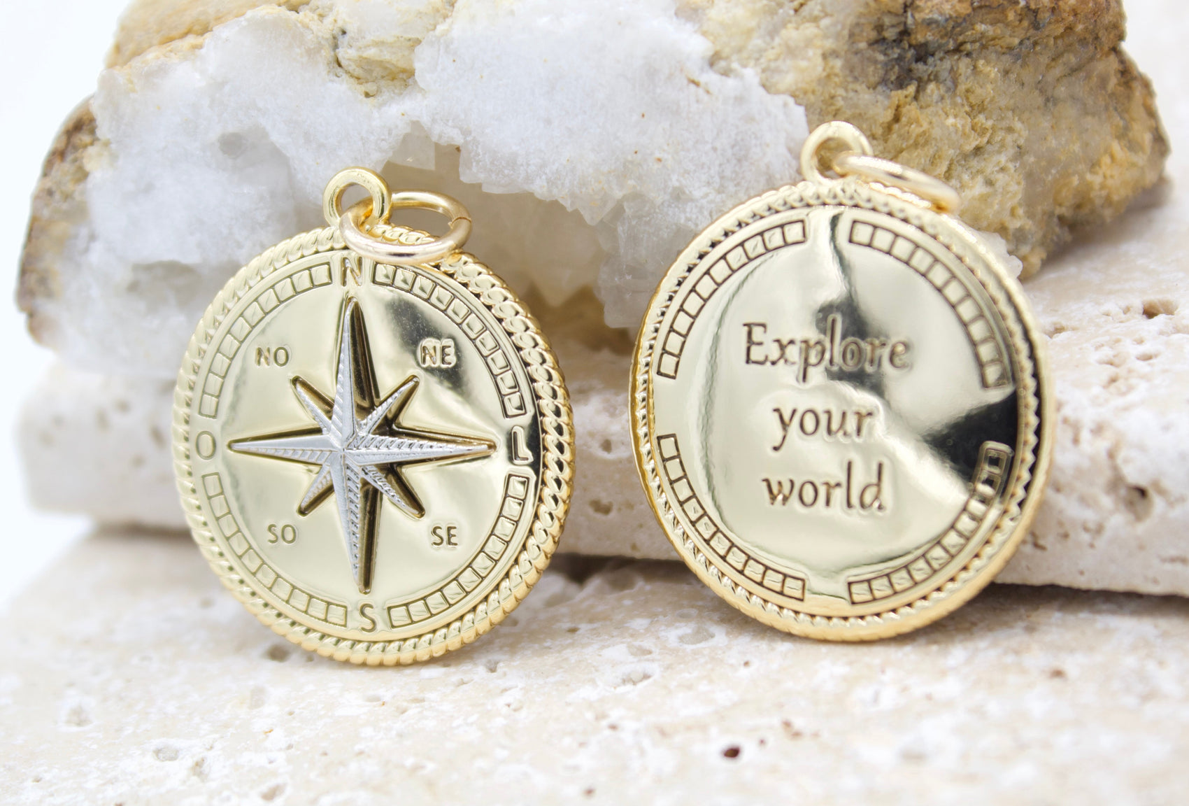Two gold compass coin pendants rest against a rough quartz crystal, displaying a wind rose on one and the inscription "Explore your world" on the other.