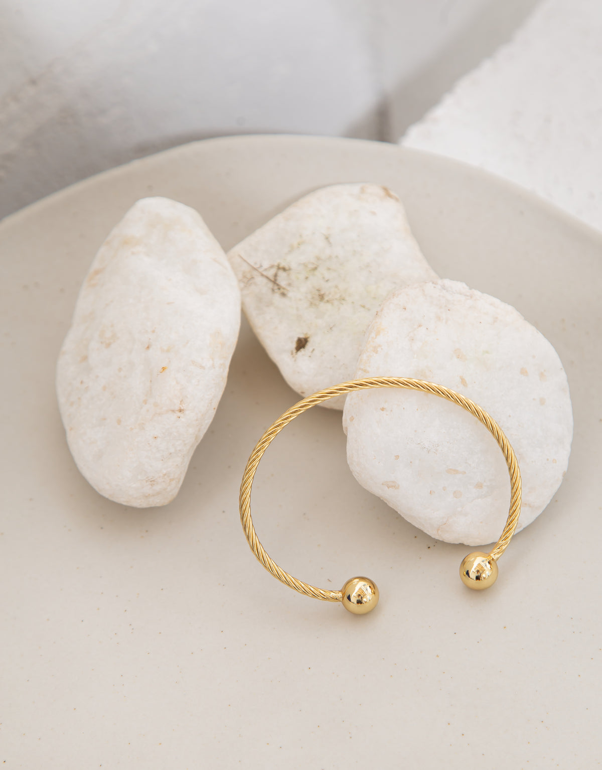A gold twisted cable cuff bracelet with polished ball ends rests on a beige tray alongside white stones.