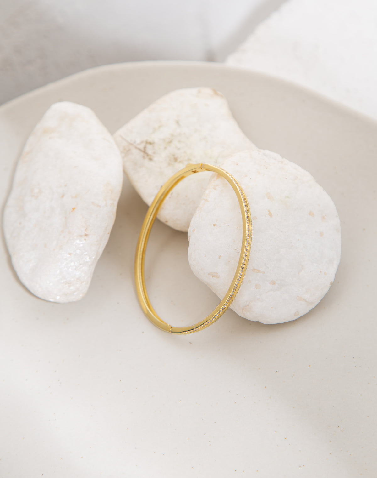 A gold pavé diamond bangle rests gracefully against smooth white decorative stones inside a neutral dish.