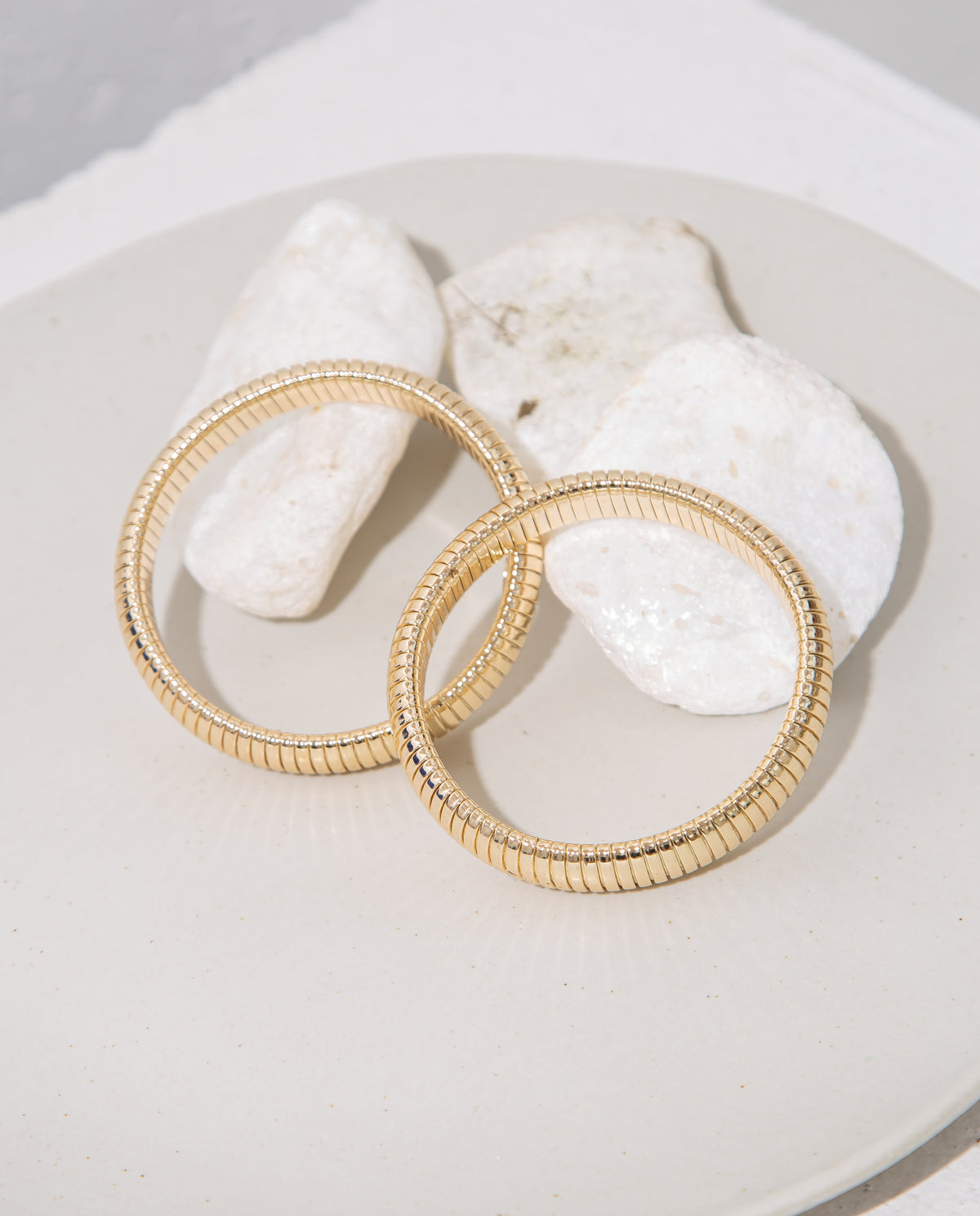 Two polished gold snake-chain bracelets rest elegantly on a white ceramic plate next to smooth white stones.