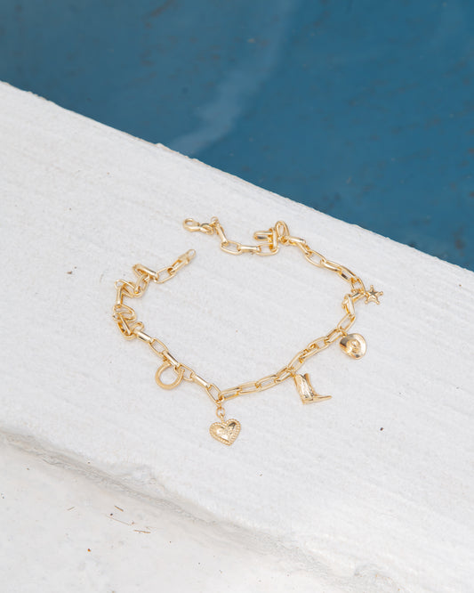A gold charm bracelet featuring western-themed pendants rests on a white ledge near a pool.