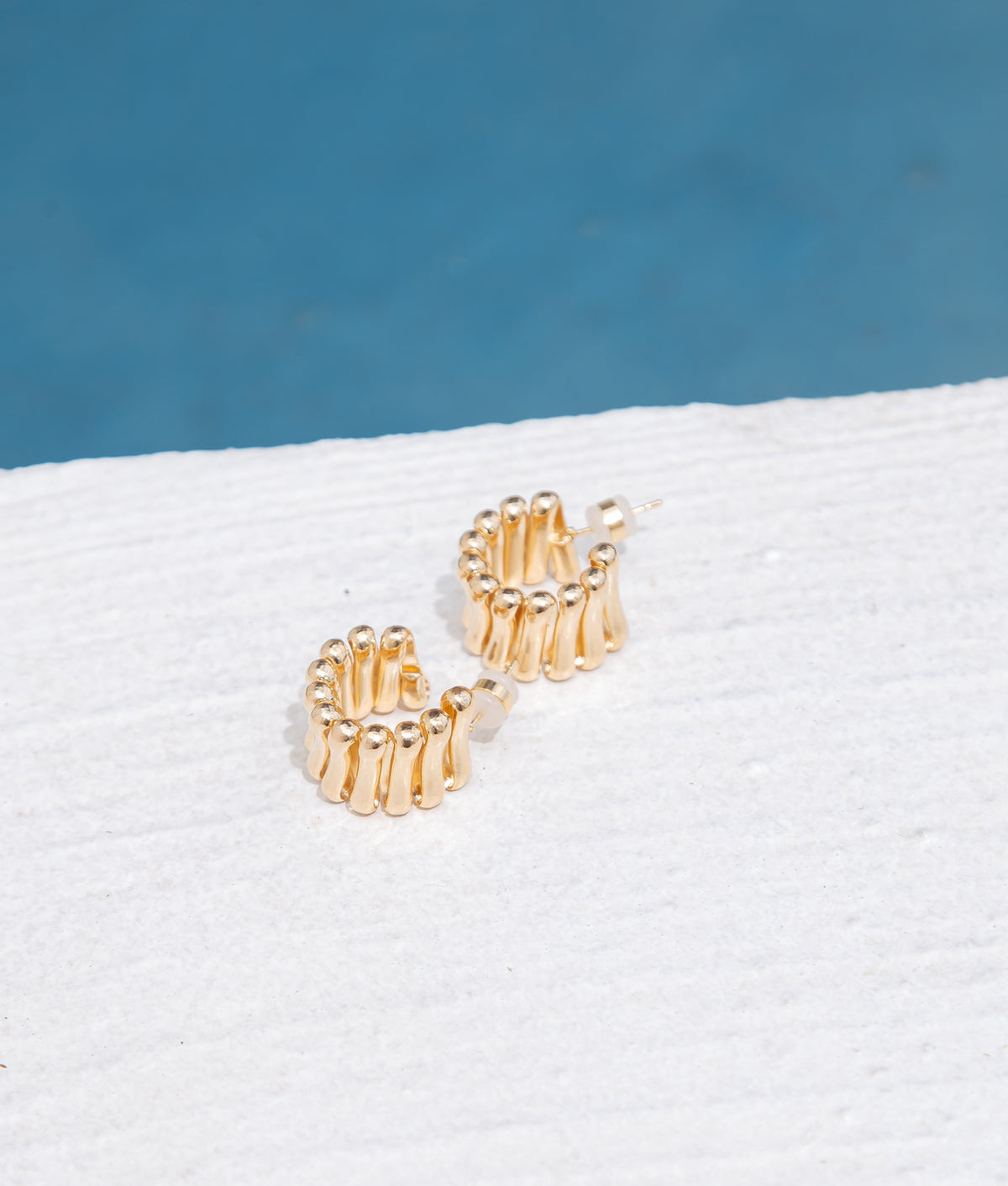 A pair of textured gold hoop earrings rests on a white surface against a blue background.