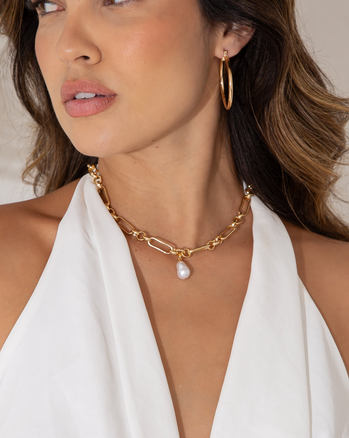 A model wears a chunky gold chain necklace with a pearl pendant and matching gold hoop earrings while wearing a white top.