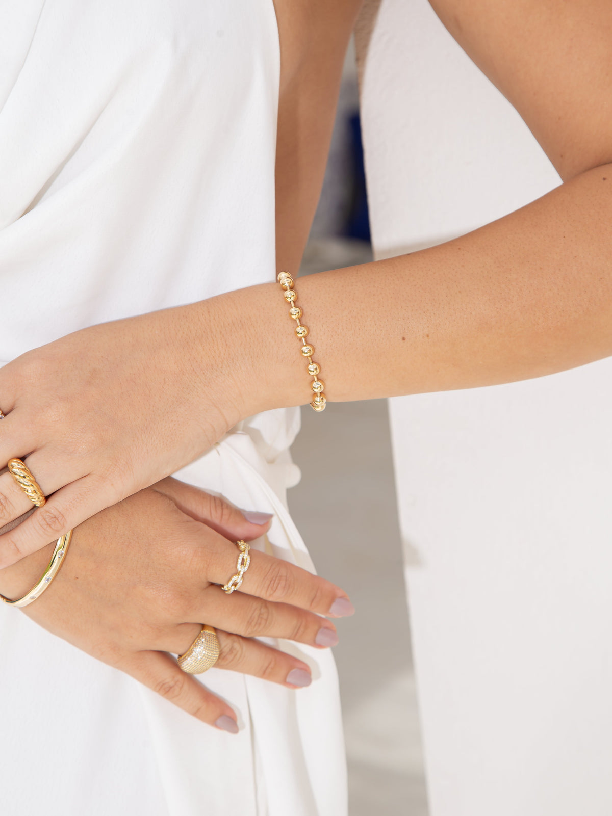 A woman wearing a gold bead bracelet and stacked rings showcases her jewelry against a white dress.