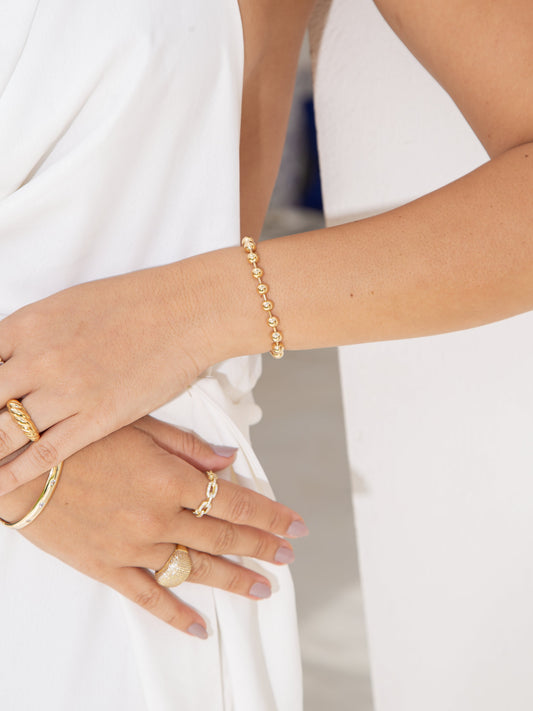 A woman wearing a gold bead bracelet and stacked rings showcases her jewelry against a white dress.