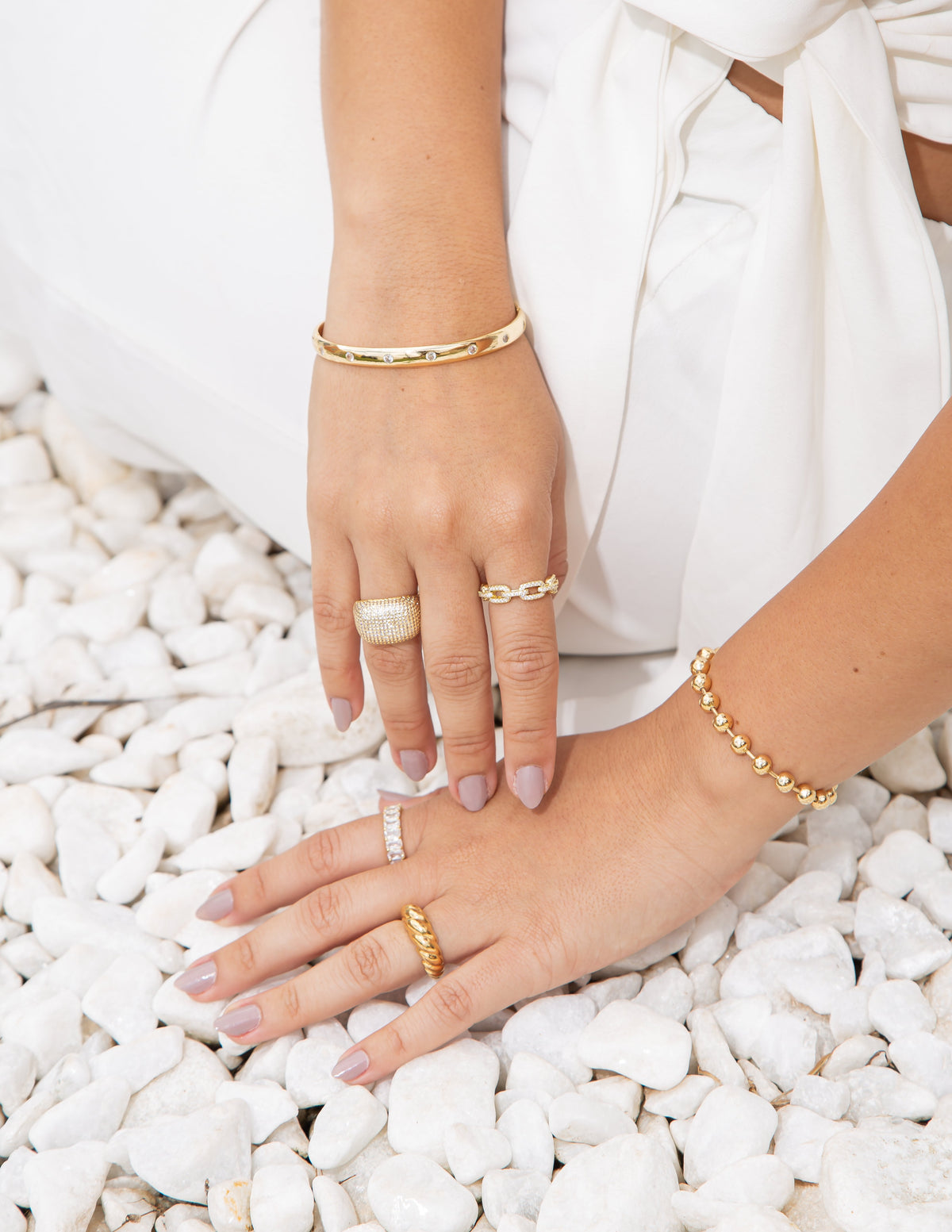 A woman wearing stylish gold jewelry, including layered rings and bracelets, rests her hands over white decorative stones.