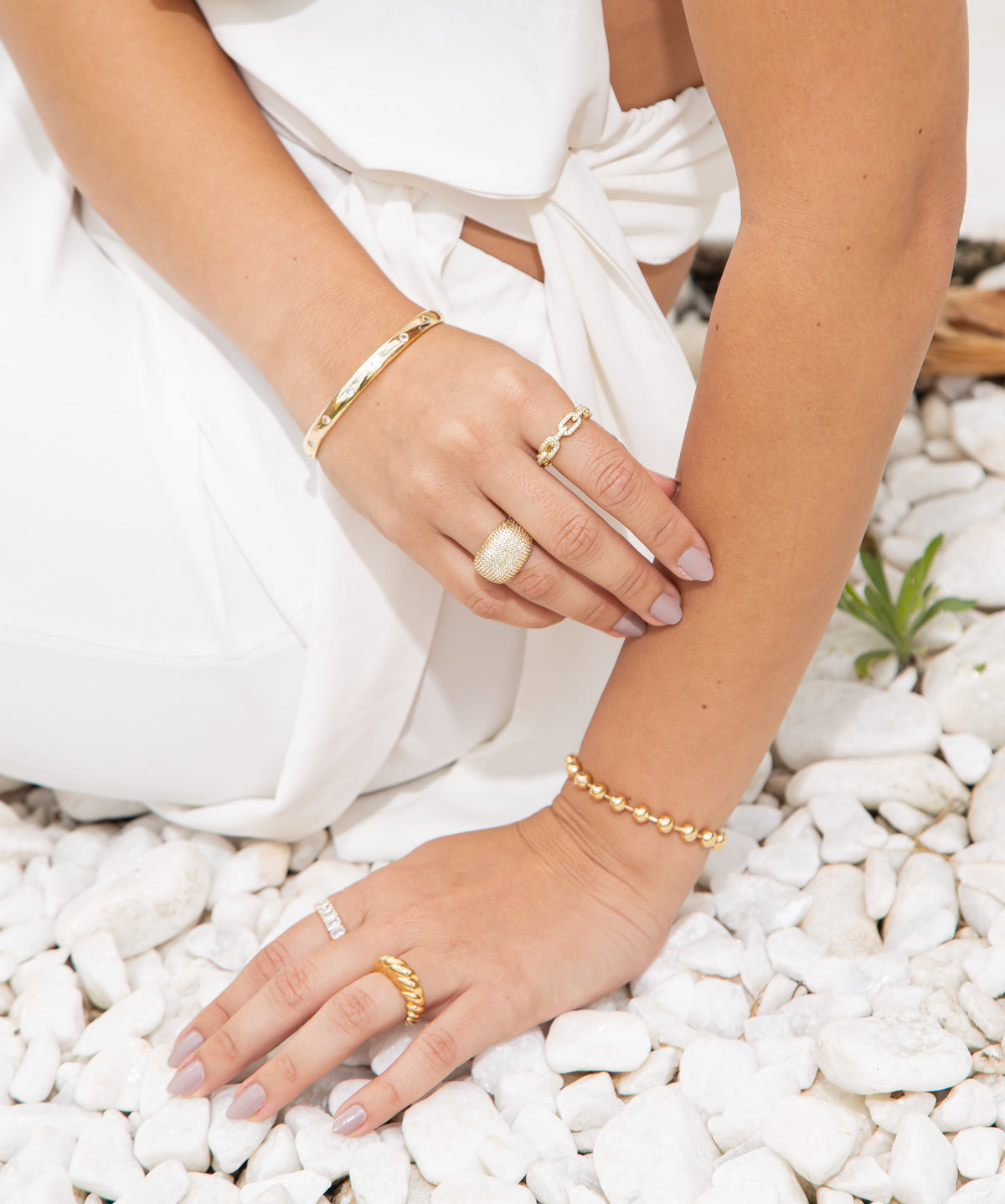 A woman wearing trendy gold jewelry, including layered rings and statement bracelets, poses gracefully over white decorative pebbles.
