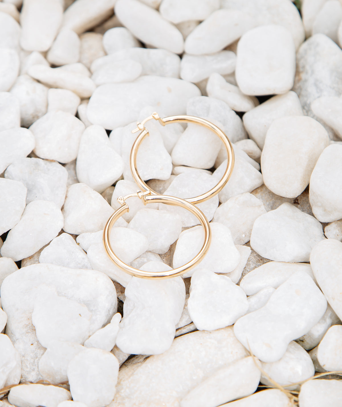 A pair of sleek gold hoop earrings rests neatly atop a bed of smooth white decorative stones.