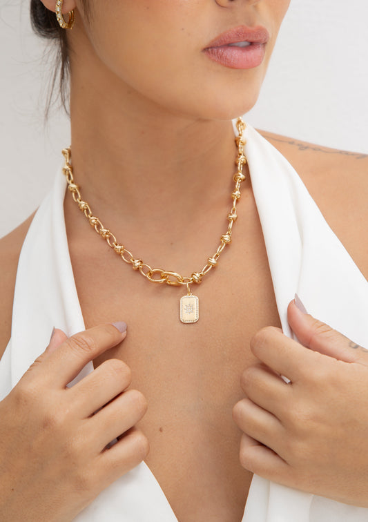 A woman wearing a gold chain necklace with a rectangular starburst pendant adjusts her white halter top.