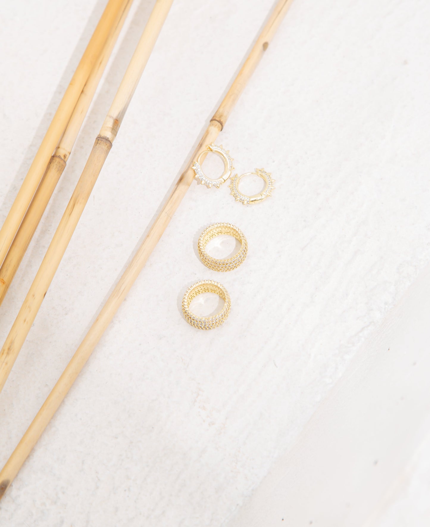 Two gold pave-set diamond huggie earrings and two matching gold eternity bands rest next to thin bamboo stalks on a light textured surface.