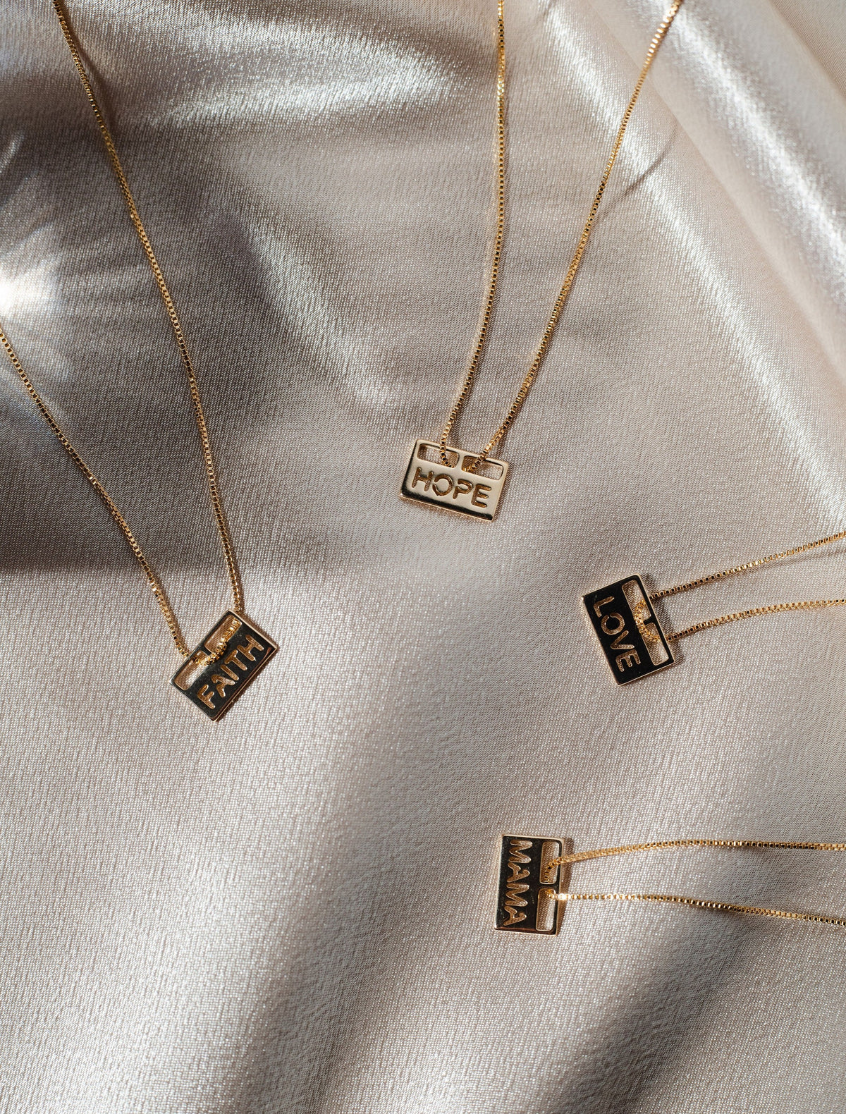 Four delicate gold bar necklaces featuring the engraved words Faith, Hope, Love, and Mama rest elegantly on a shimmering satin background.