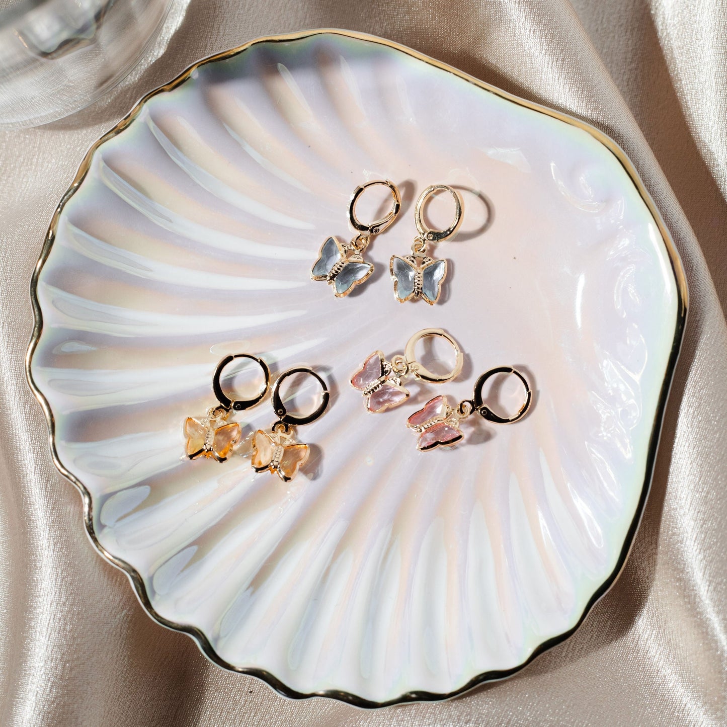 Three pairs of gold hoop earrings with colorful butterfly charms rest gracefully inside an iridescent shell-shaped dish.