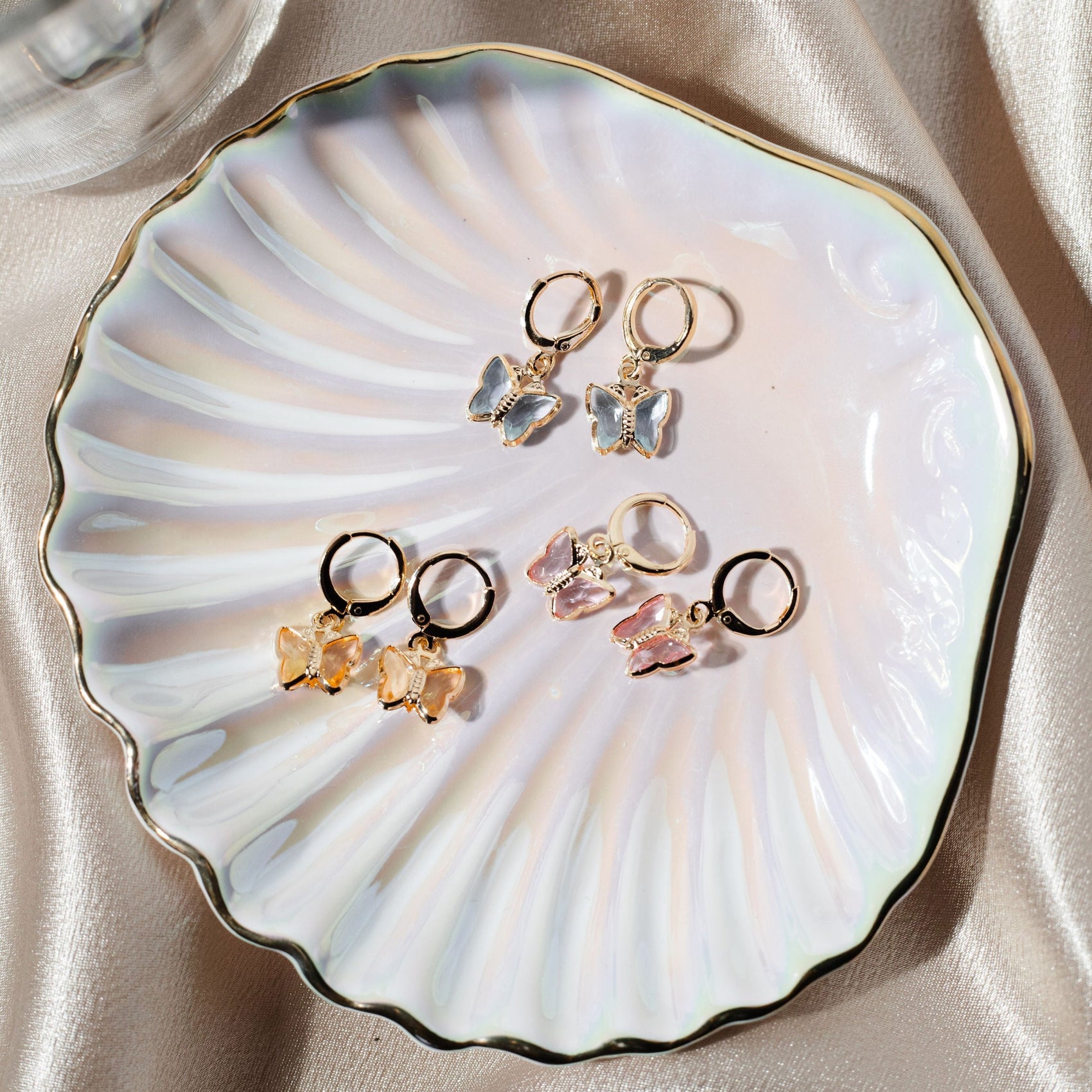 Three pairs of gold hoop earrings with colorful butterfly charms rest gracefully inside an iridescent shell-shaped dish.