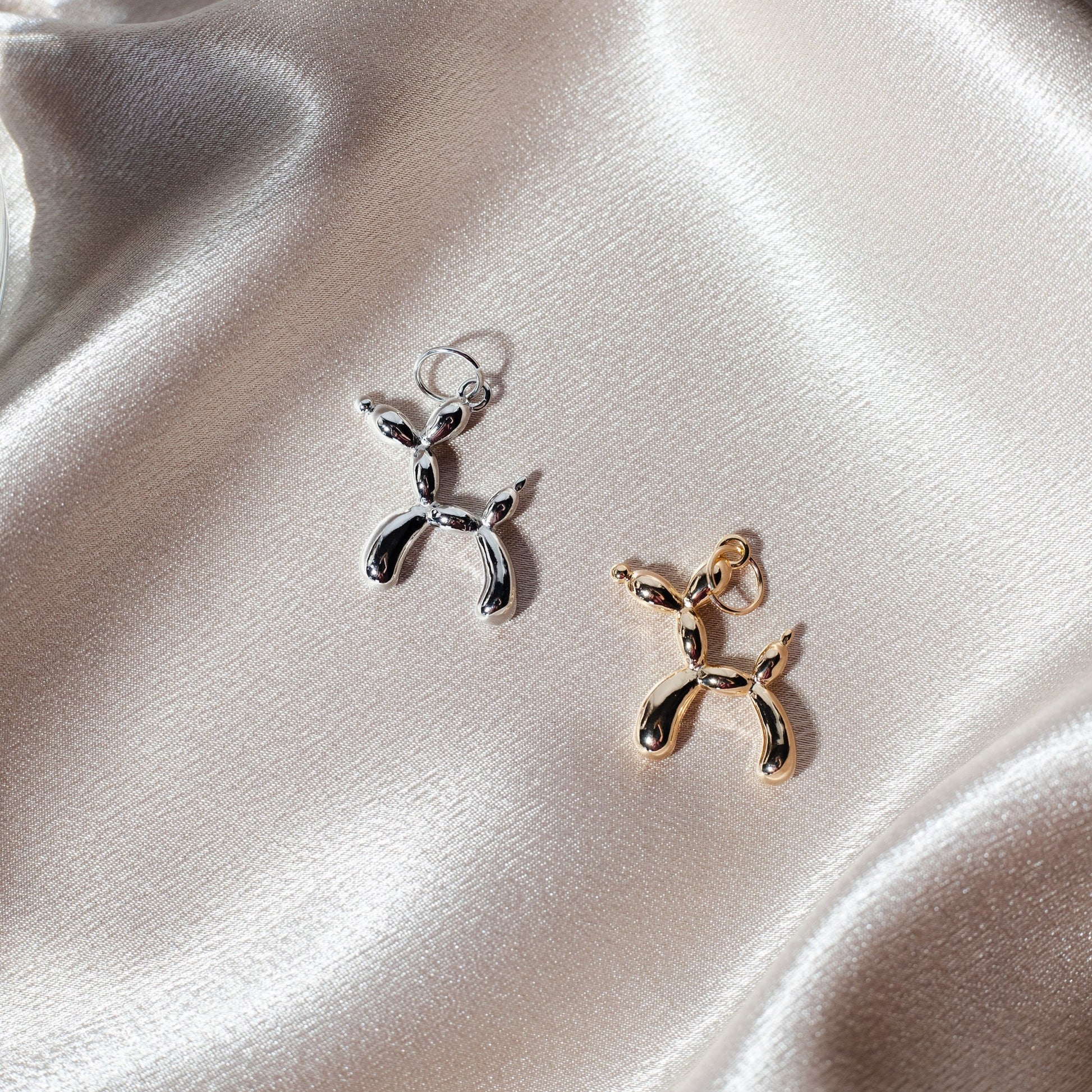 A silver and a gold balloon dog charm rest on a silky, neutral-toned fabric background.