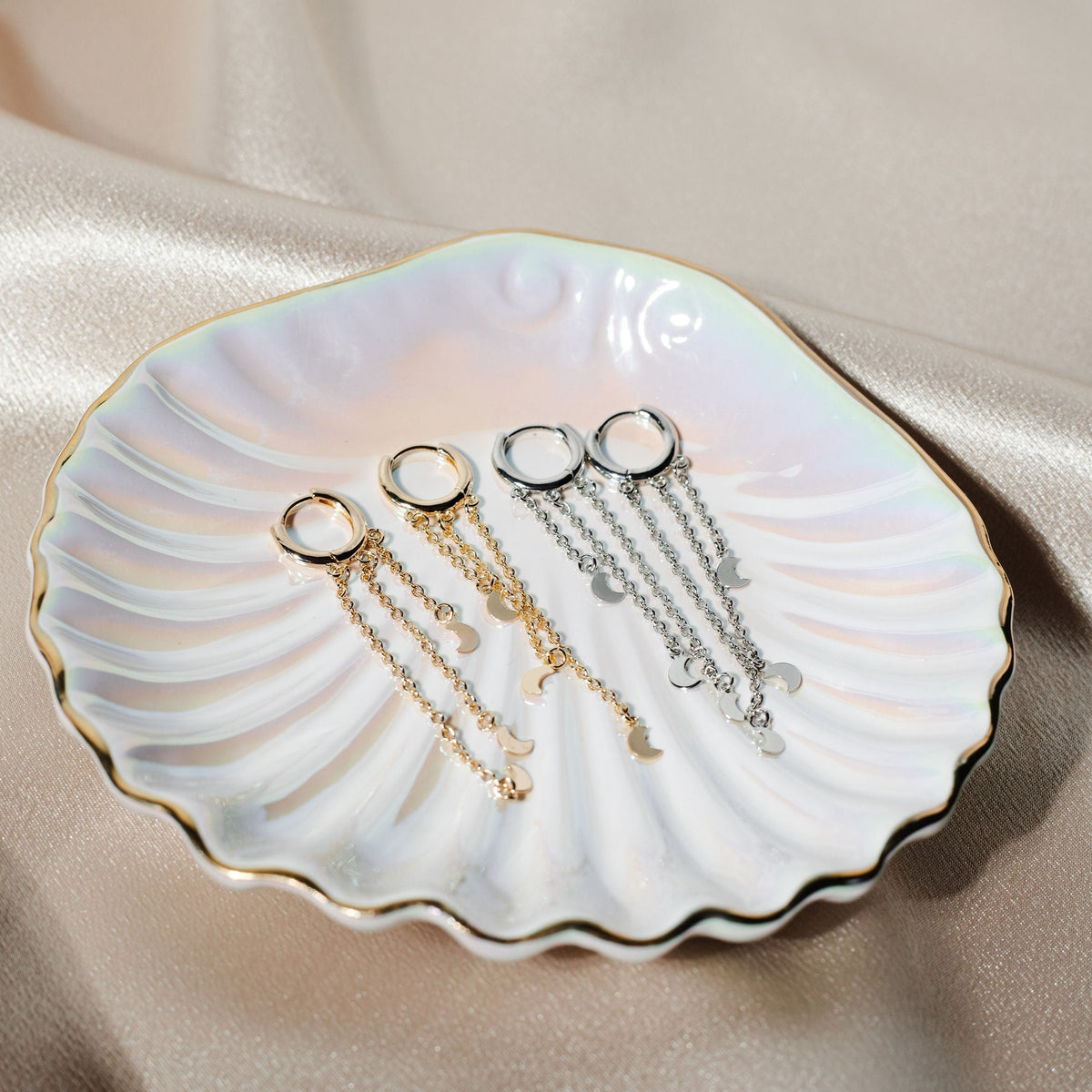 Two pairs of gold and silver dangle hoop earrings with delicate crescent moon charms rest neatly inside an iridescent, shell-shaped jewelry dish on a silky fabric backdrop.