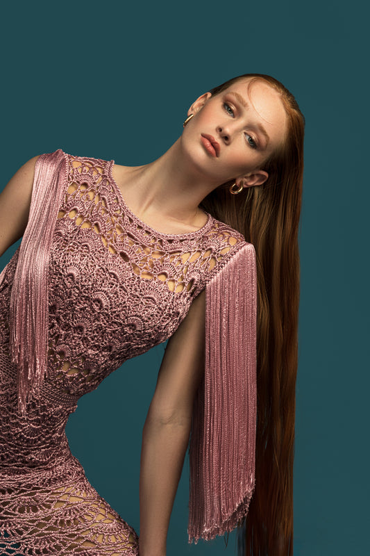 A red-haired model wearing a pink crochet dress with fringe details poses against a teal backdrop.