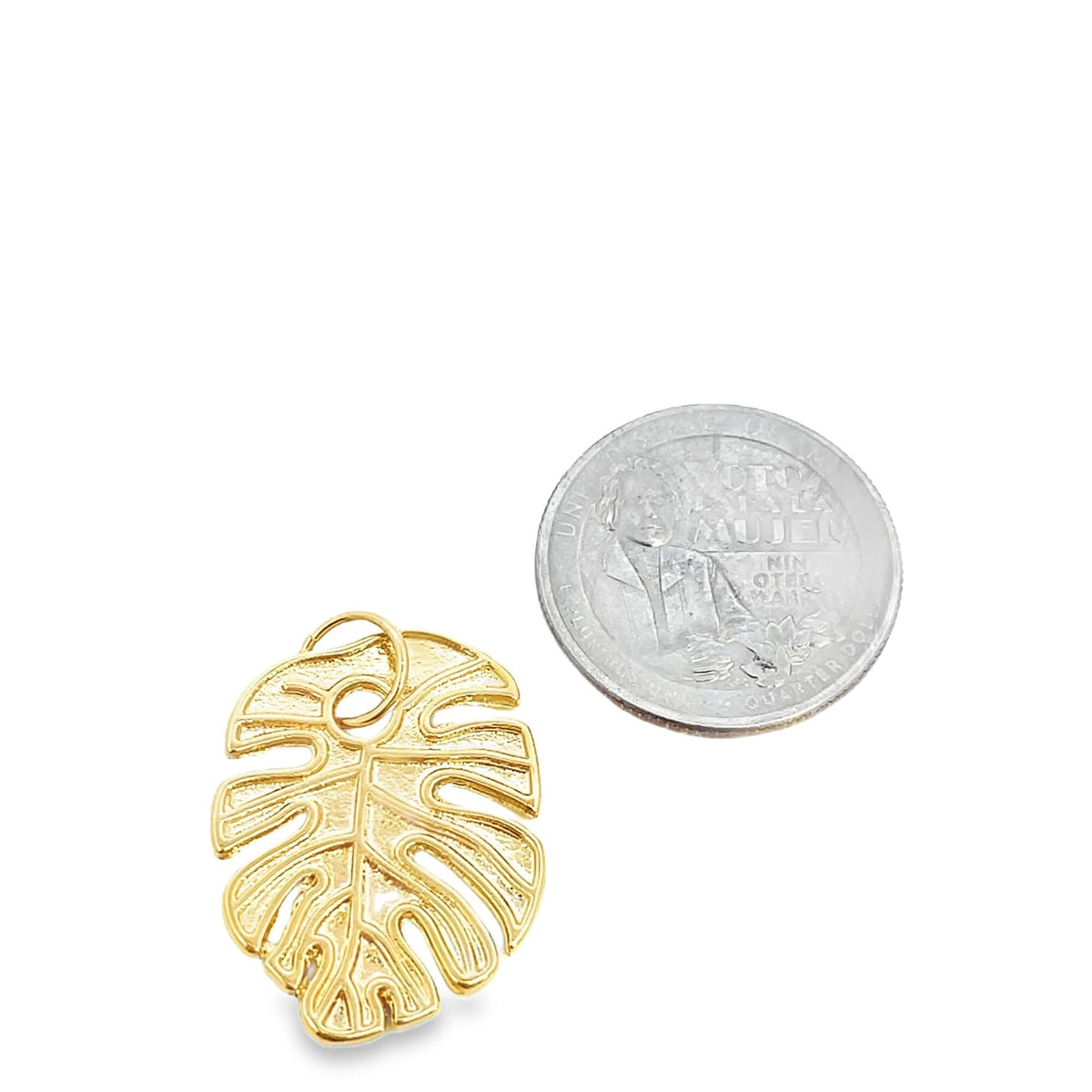 A gold monstera leaf charm rests beside a US quarter for size comparison on a white background.