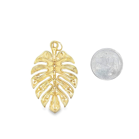 A shiny gold monstera leaf pendant rests next to a US quarter coin for scale on a white background.