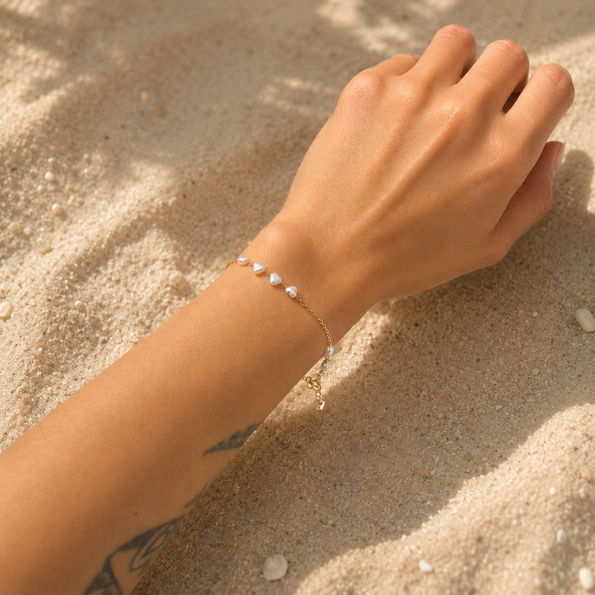 A woman wears a delicate gold chain bracelet featuring small white heart-shaped pearls resting on a sandy beach.