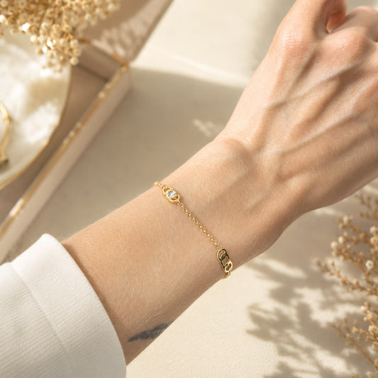 A delicate gold chain bracelet featuring a sparkling gemstone and letter charms rests on a woman's wrist.