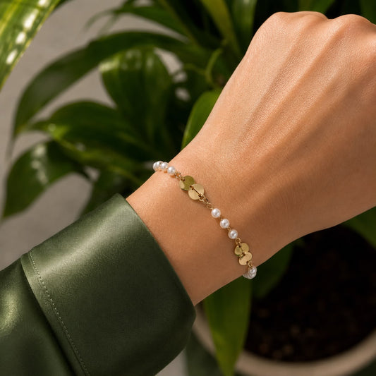 A delicate gold chain bracelet featuring alternating pearls and small circular discs adorns a wrist against a backdrop of lush green leaves.