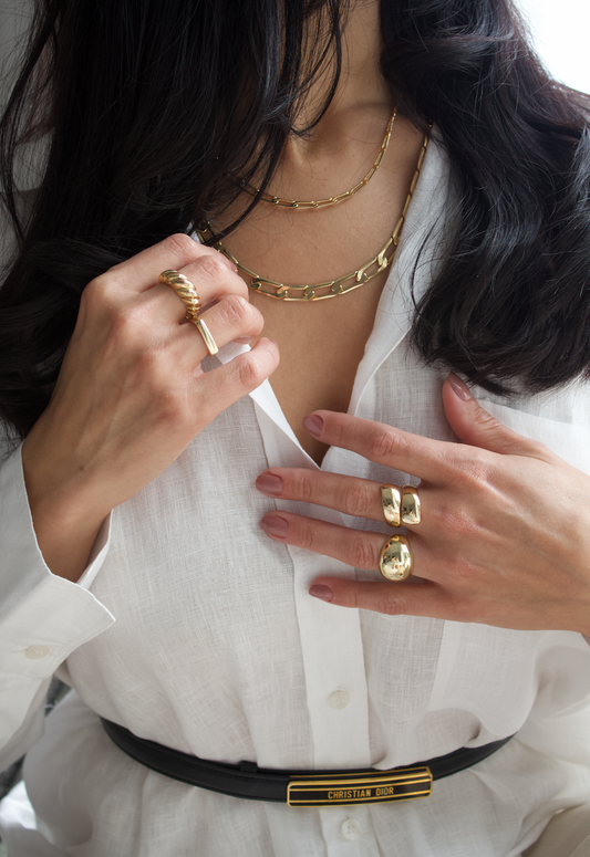 A woman wearing layered gold chain necklaces and statement gold rings poses in a crisp white shirt.
