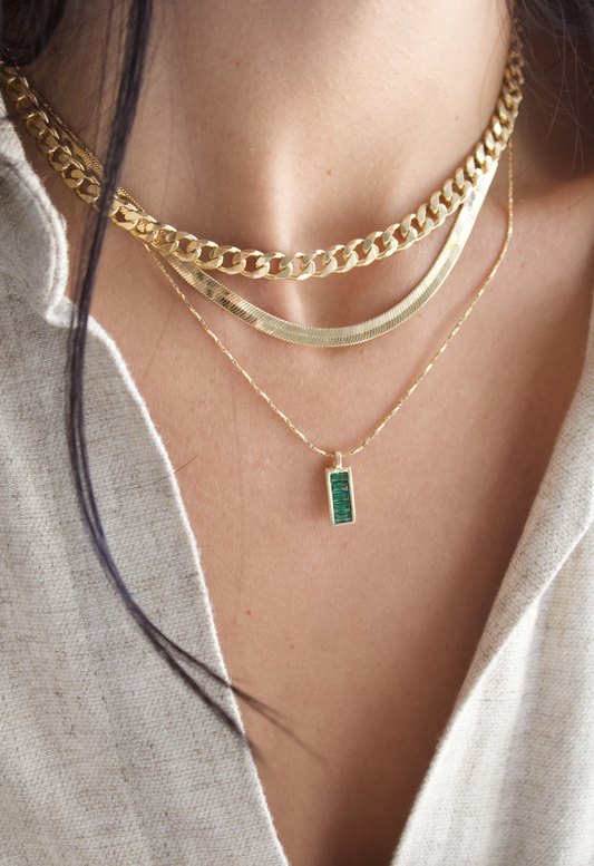 A woman wearing a layered gold necklace set featuring a chunky chain, a flat herringbone chain, and a delicate pendant with a rectangular green gemstone.