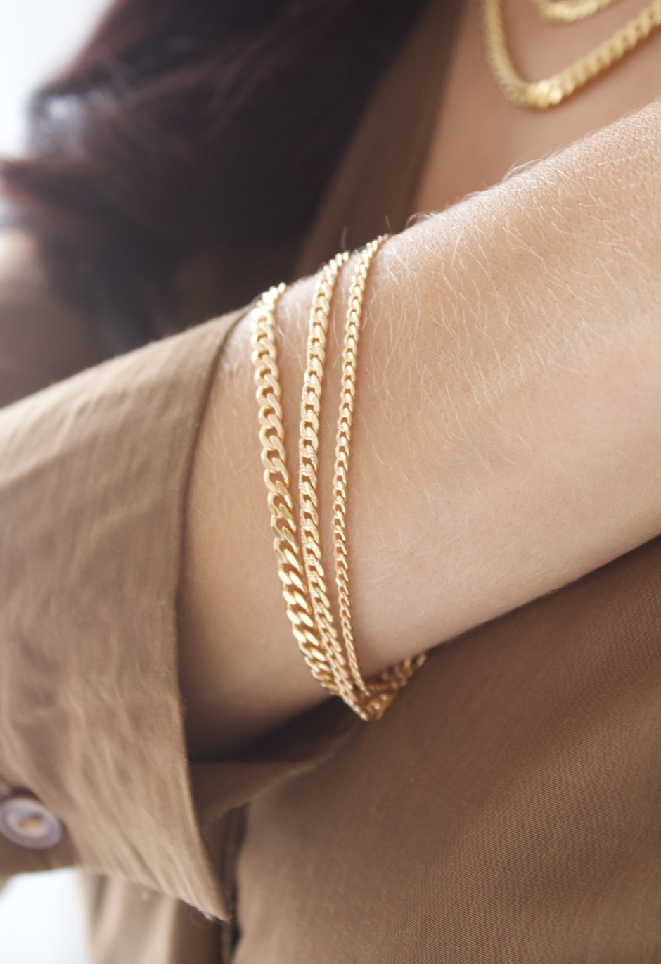 A woman wears a stack of delicate gold chain bracelets on her forearm while dressed in a brown blouse.