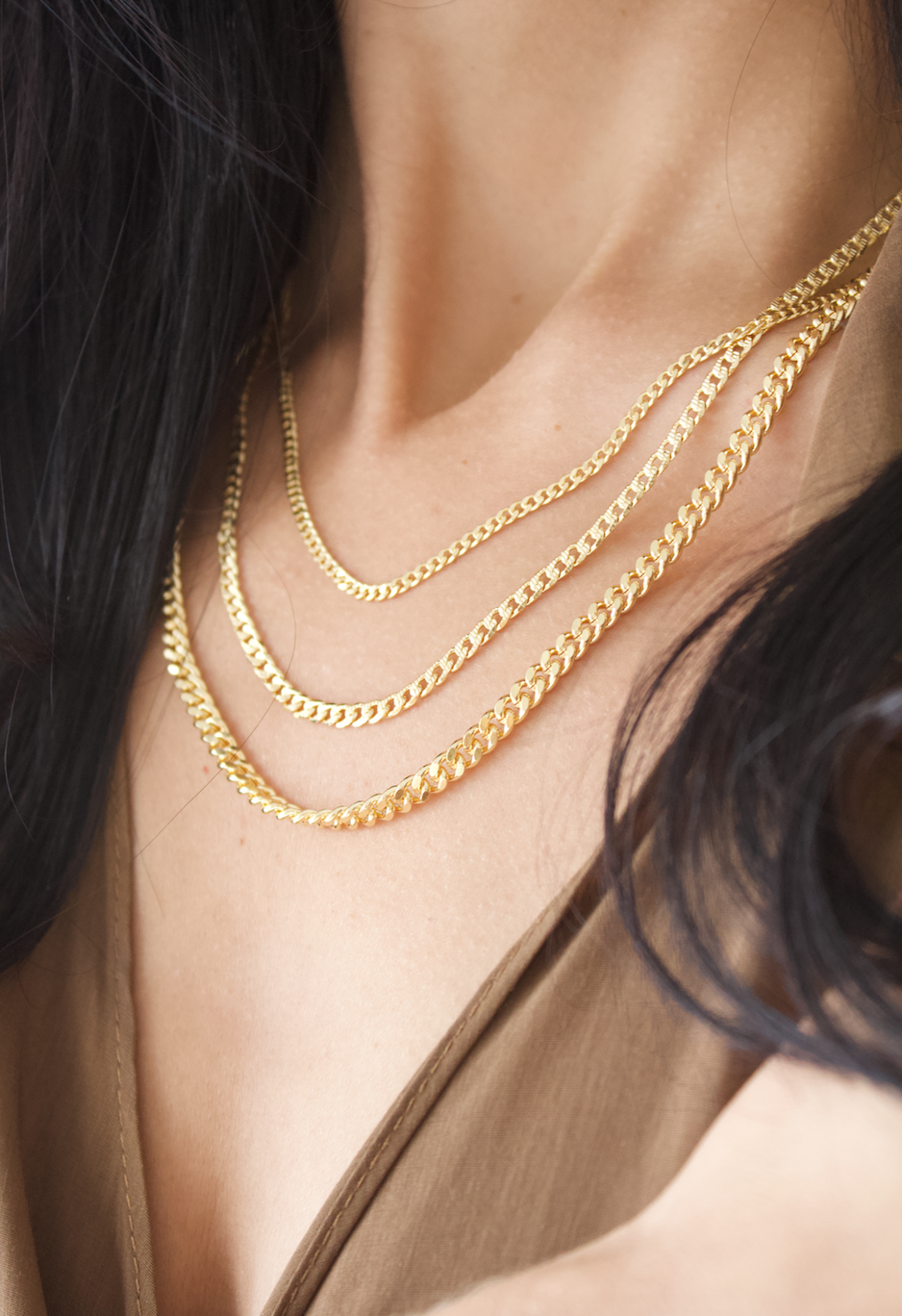 A woman with dark hair wears three layered gold cuban link necklaces against a neutral-toned top.
