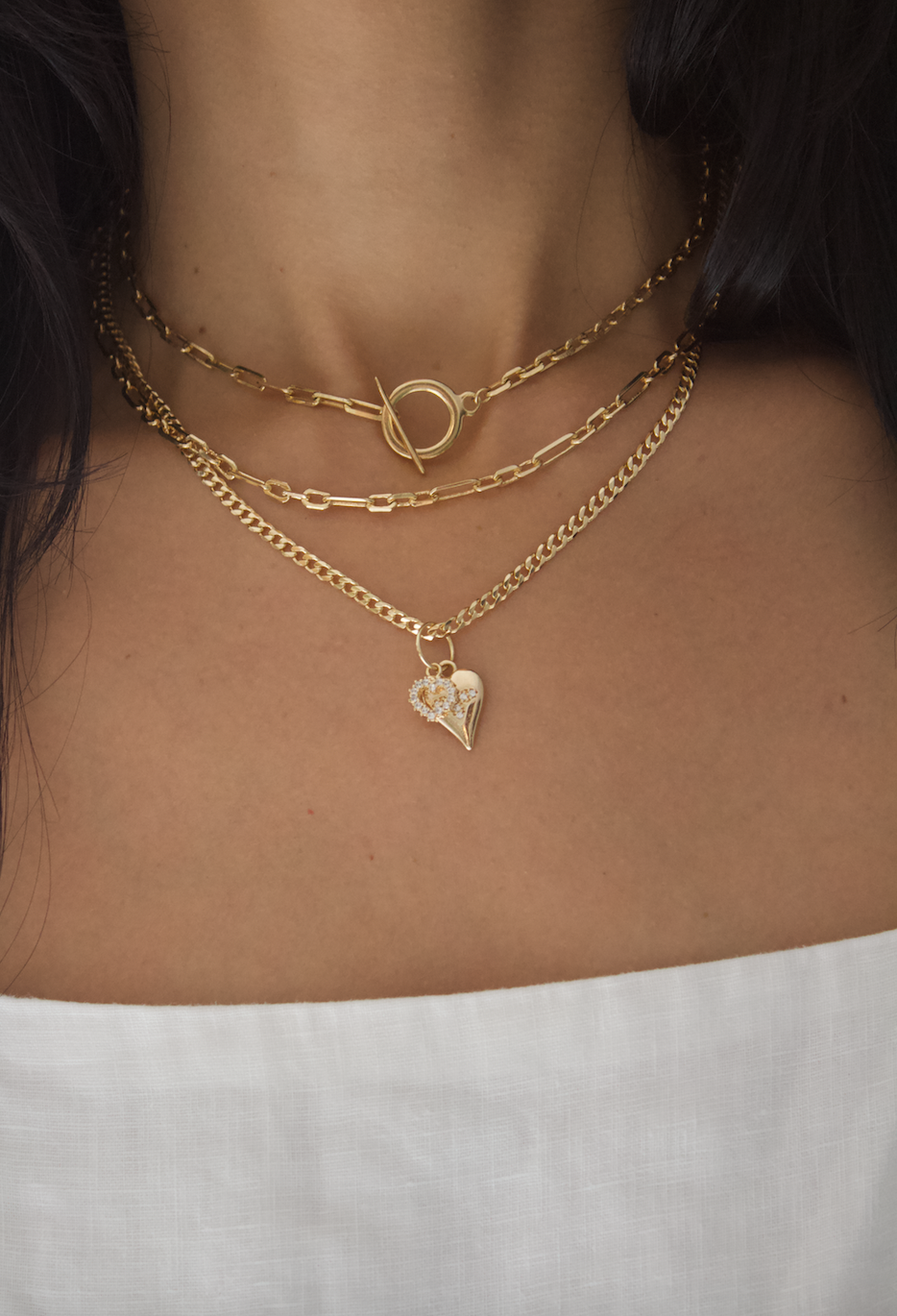 A woman wears three layered gold chain necklaces, featuring a toggle clasp and a sparkling heart pendant.