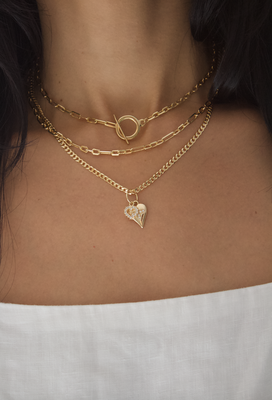 A woman wears three layered gold chain necklaces, featuring a toggle clasp and a sparkling heart pendant.