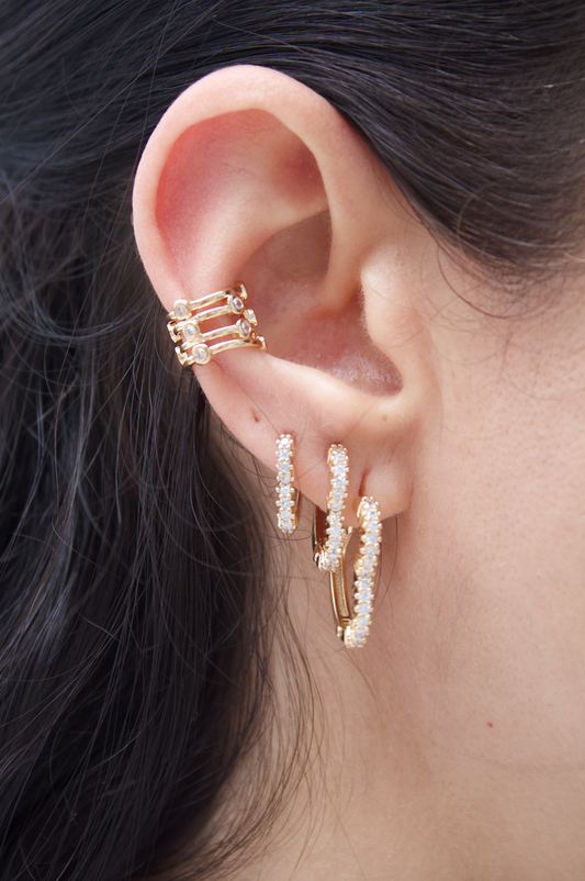 A person's ear features a multi-band gold cartilage cuff and three sparkling pave diamond huggie hoop earrings.