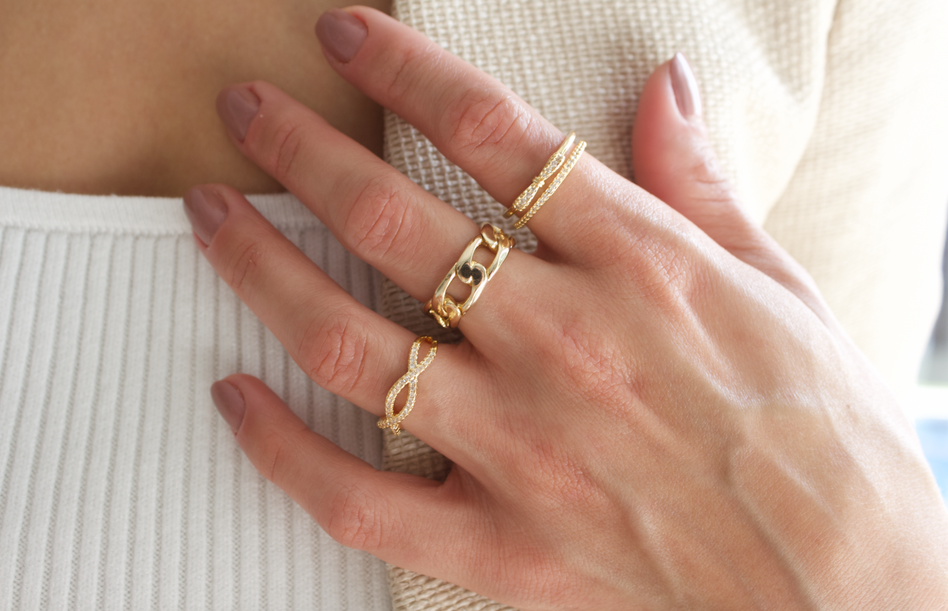 A woman wears a collection of elegant gold rings on her fingers, showcasing a chunky chain-link ring and delicate diamond-accented bands.