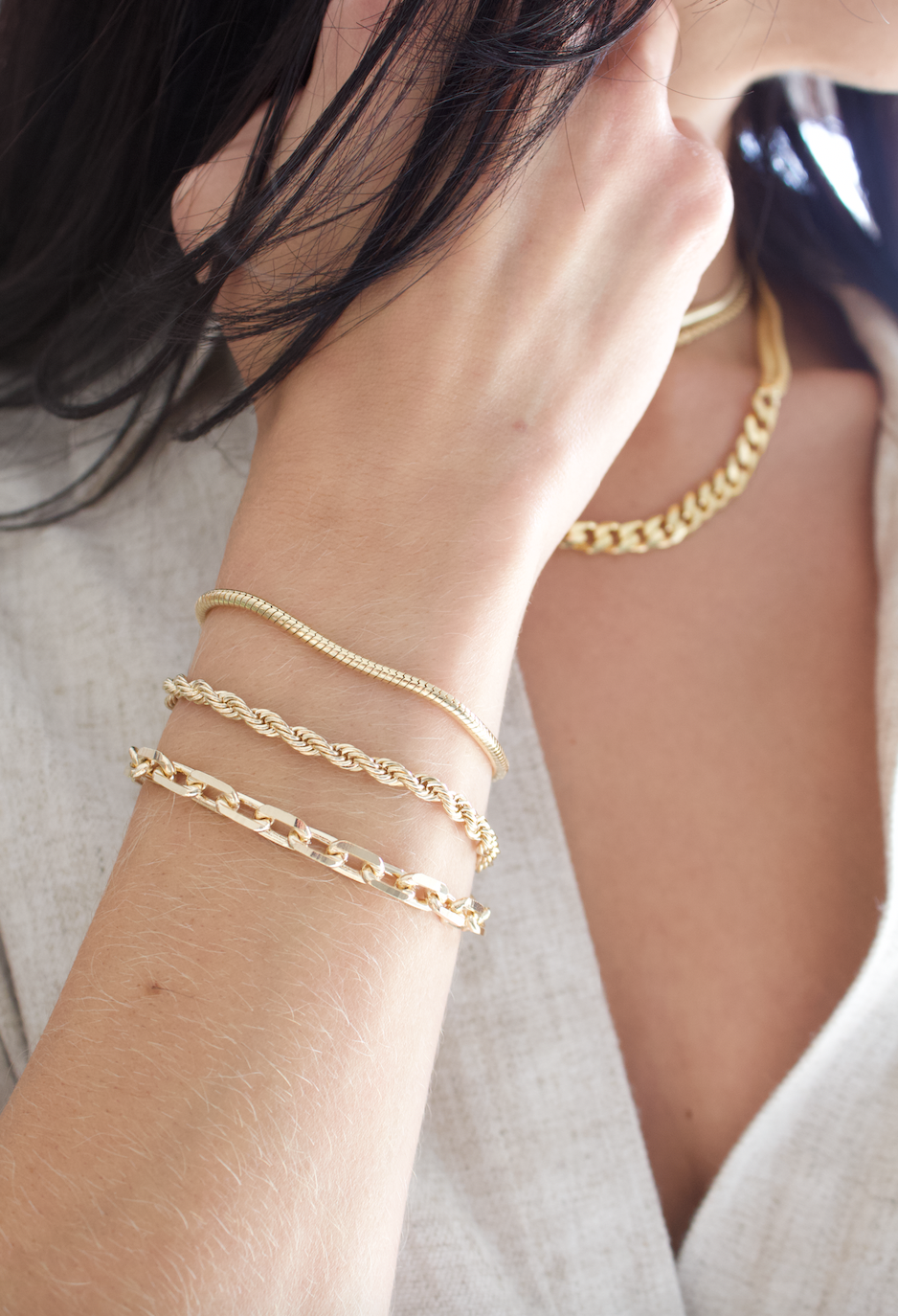 A stylish woman showcases a stack of three delicate gold bracelets on her arm while wearing matching gold necklaces.