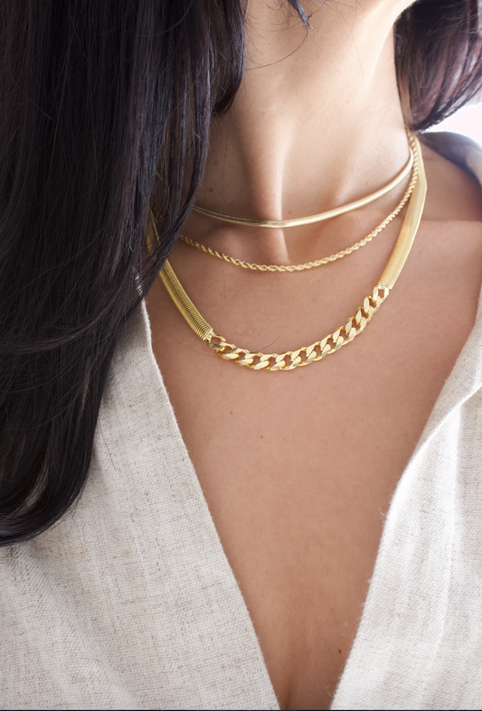 A woman wears a layered set of gold necklaces, featuring a sleek herringbone chain, a delicate rope chain, and a bold curb link necklace against a cream-colored top.