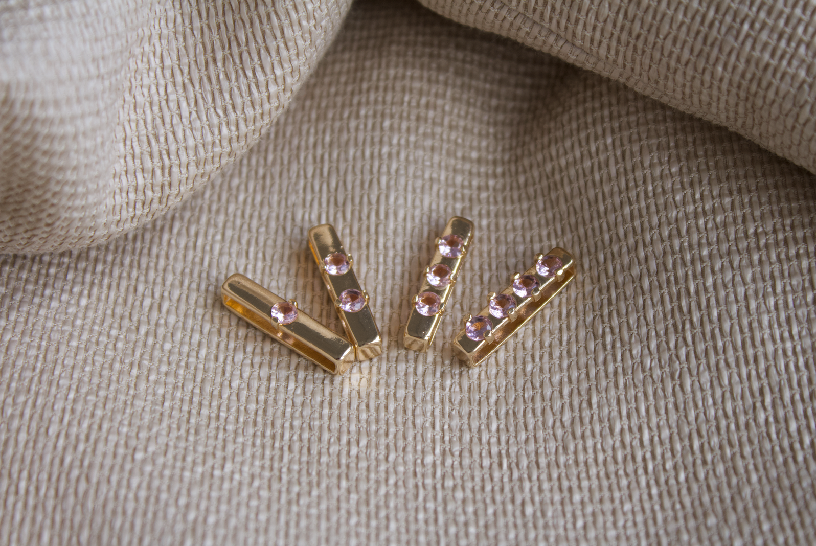Four gold slider charms with inset pink crystals rest on a textured beige fabric background.